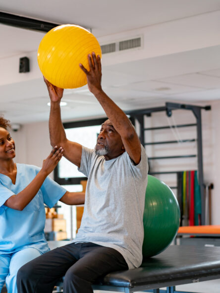 Cheerful physiotherapist helping senior black patient workout with a fitness ball at a rehabilitation center  - healthcare concepts